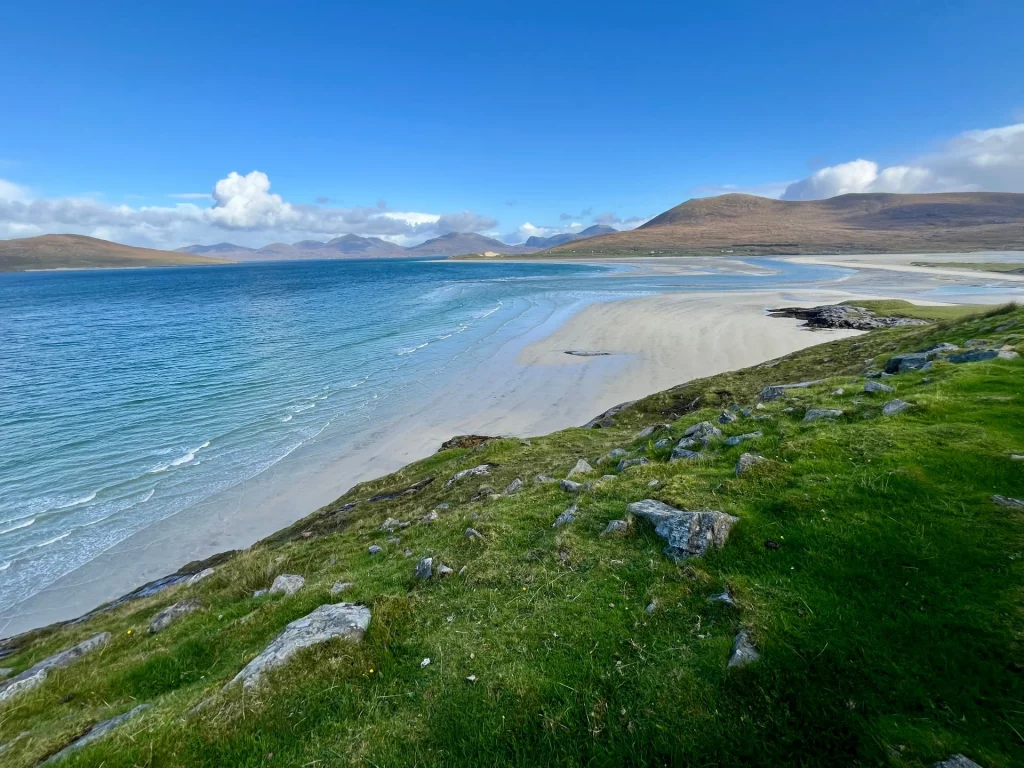 Blick von einem grasbewachsenen, felsigen Hang hinunter auf einen weiten, weißen Sandstrand und türkisfarbenes Meer. Im Hintergrund runden sanfte, braun-grüne Hügelketten die maritime Landschaft ab.
