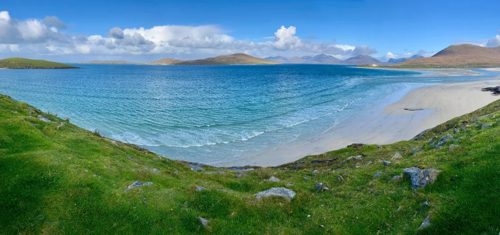 Eine weite Panorama-Aufnahme einer halbmondförmigen Bucht mit strahlend weißem Sandstrand und türkisfarbenem Wasser. Im Vordergrund blickt man von einem grünen, grasbewachsenen Hügel hinab. Im Hintergrund erstreckt sich eine sanfte, bräunliche Bergkette unter einem strahlend blauen Himmel mit vereinzelten weißen Wolken.