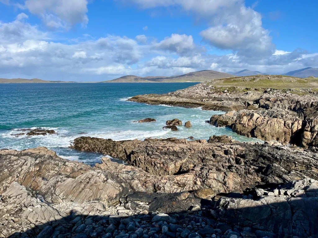 Eine zerklüftete, graue Felsküste, an der sich weiße Gischt im dunkelblauen Meer bricht. Im Hintergrund sind sanfte Hügel unter einem leicht bewölkten Himmel zu sehen.