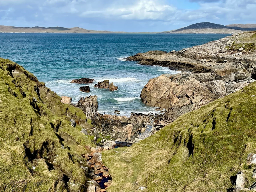 Blick durch eine grüne Senke auf eine felsige Bucht. Das türkisfarbene Wasser schäumt zwischen den Steinen, während am Horizont weitere Inselketten und Hügel sichtbar sind.