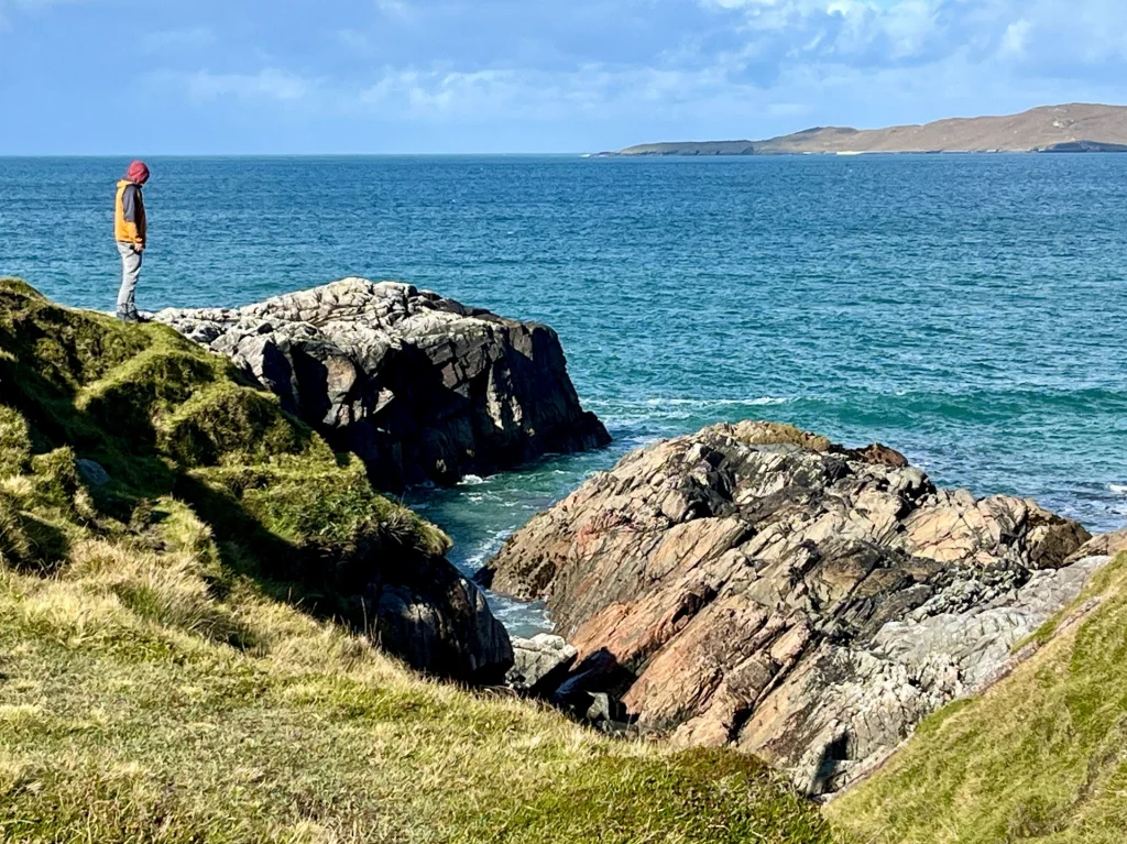 Eine Person in einer orangen Jacke steht am Rand einer begrünten Klippe und blickt auf eine schmale Meeresbucht hinunter, die von steilen Felswänden flankiert wird.