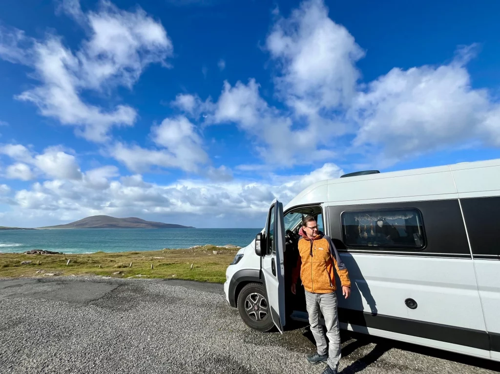 Ein hellgrauer Kastenwagen parkt auf einem Schotterplatz direkt am Meer. Ein Mann in einer orangefarbenen Jacke lehnt lächelnd an der offenen Fahrertür, im Hintergrund glitzert das Wasser unter strahlend blauem Himmel.