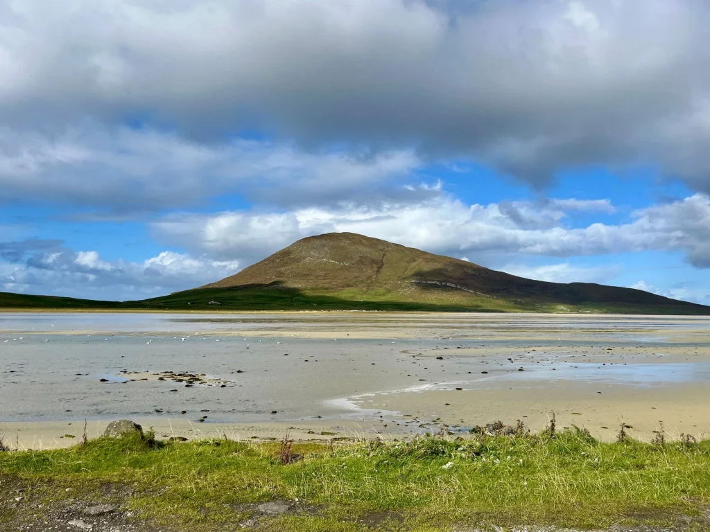 Eine weite, flache Sandbucht bei Ebbe mit vereinzelten Wasserpfützen. Im Hintergrund erhebt sich ein markanter, grüner Hügel unter einer dramatischen Wolkendecke.
