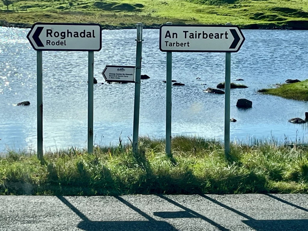 Zweisprachige Straßenschilder (Gälisch/Englisch) an einer Uferstraße, die links nach Rodel (Roghadal) und rechts nach Tarbert (An Tairbeart) weisen.