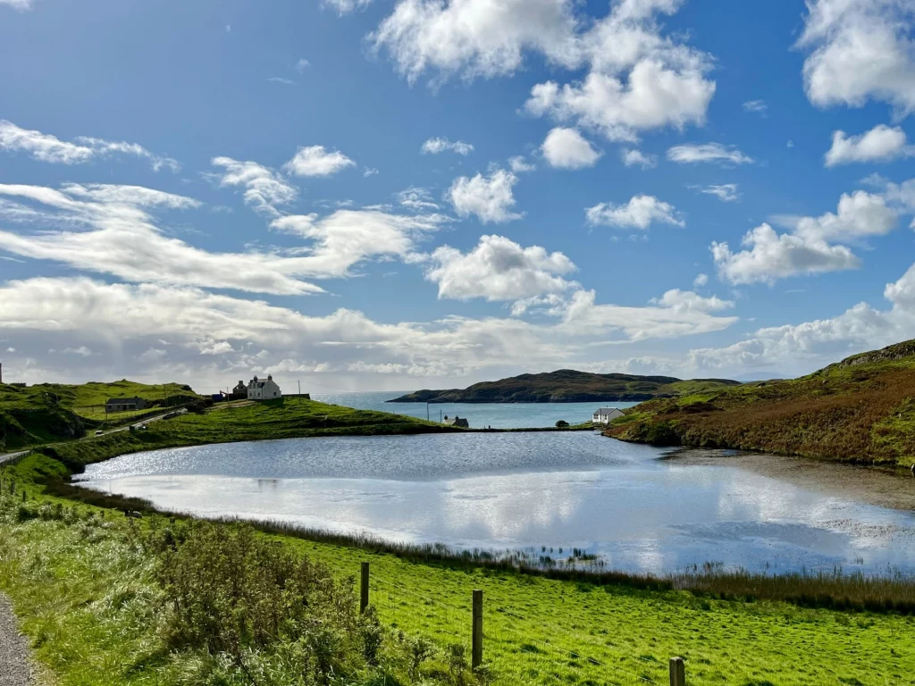 Weite Landschaft auf der Isle of Harris mit einer ruhigen Meeresbucht unter einem strahlend blauen Himmel mit Schäfchenwolken.