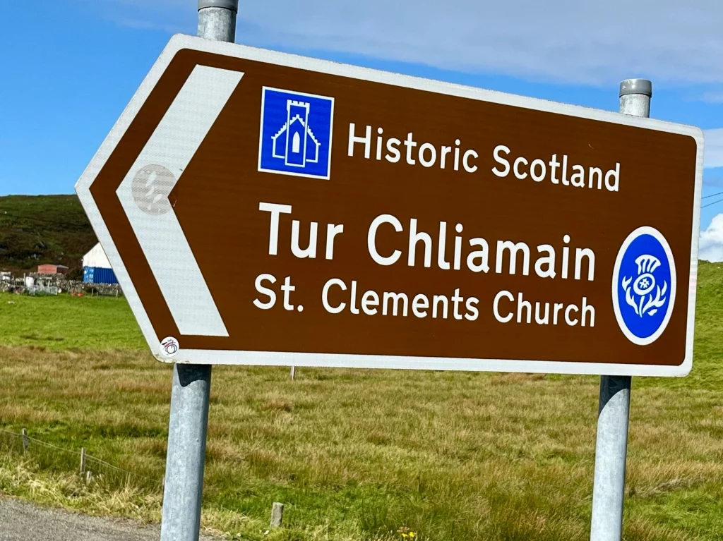 Ein braunes schottisches Touristenschild mit der Aufschrift „Historic Scotland – Tur Chliamain – St. Clement’s Church“. Das Schild zeigt zudem ein Piktogramm einer Kirche und ein blaues Siegel.