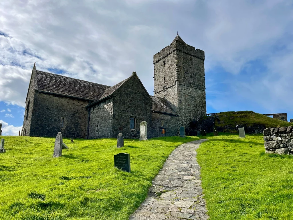 Eine imposante, mittelalterliche Steinkirche (St. Clement's Church) mit einem massiven quadratischen Turm unter einem weiten, leicht bewölkten Himmel. Ein schmaler Steinpfad führt durch den grünen Kirchhof mit vereinzelten, alten Grabsteinen direkt zum Gebäude.