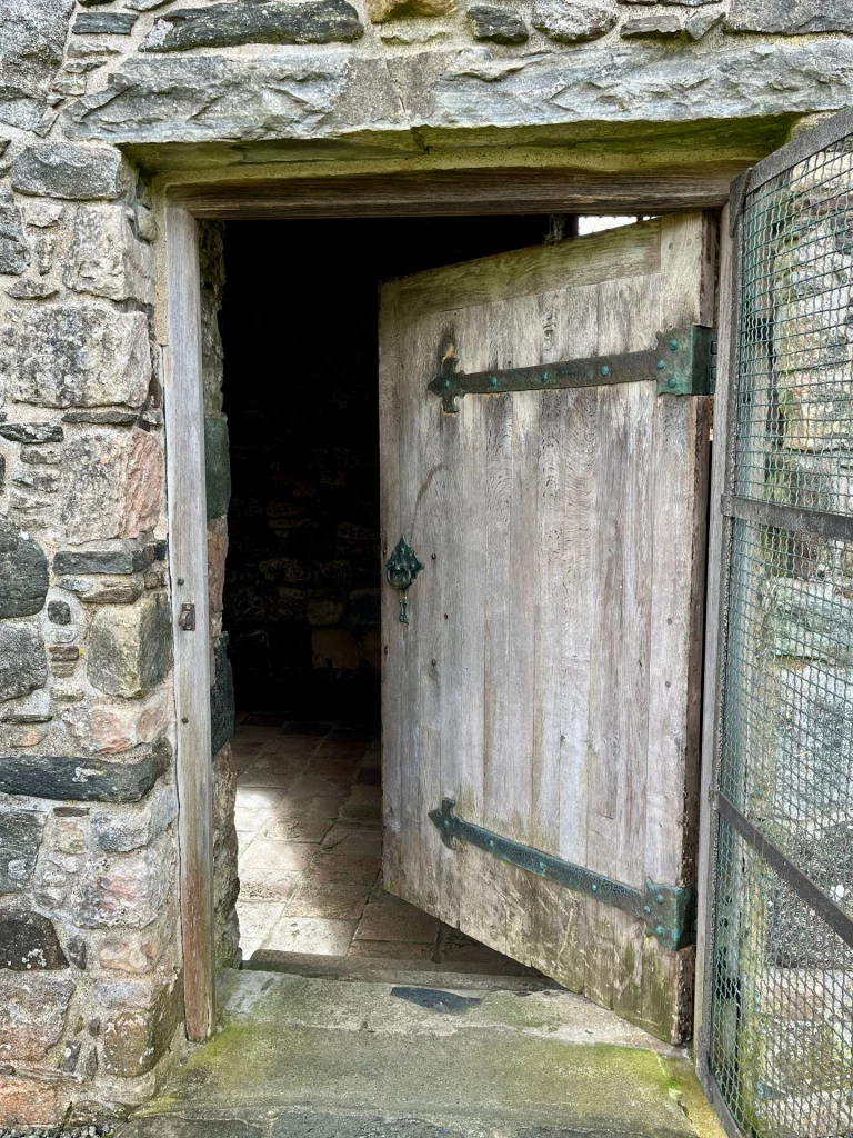 Eine schwere, rustikale Holztür mit Eisenbeschlägen steht in einem massiven Steinrahmen offen und gibt den Blick in das kühle, schattige Innere der Kapelle frei.
