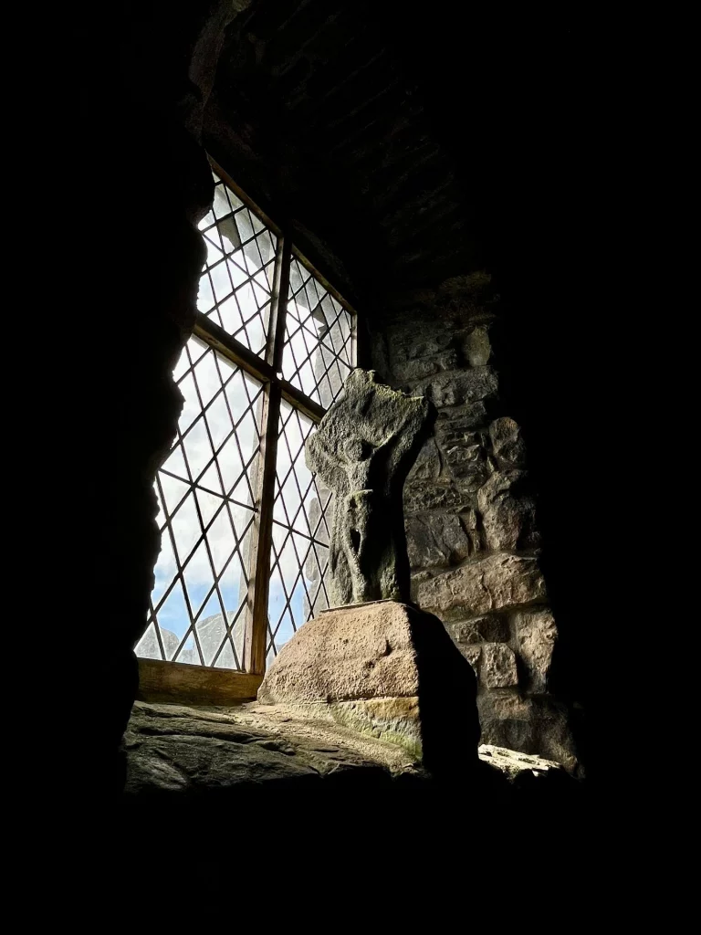 Ein kleines, bleigefasstes Rautenfenster in einer tiefen Mauernische, durch das helles Tageslicht in den ansonsten dunklen Kirchenraum fällt.