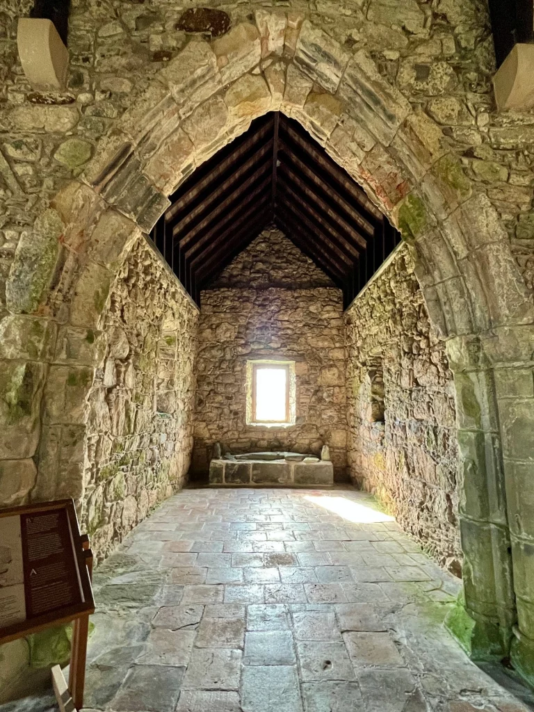 Blick durch einen spitz zulaufenden Steinbogen in das schmale, leere Kirchenschiff der St. Clement’s Church mit rauen Steinwänden und einem tief hängenden Holzdach.