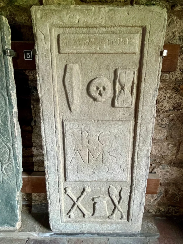 Eine rechteckige Grabplatte aus dem Jahr 1725 mit markanten Memento-mori-Symbolen. Im oberen Bereich sind ein Sarg, ein Totenkopf und eine Sanduhr eingraviert. Darunter befinden sich die Initialen „RC AMS“ und das Datum. Den unteren Abschluss bilden gekreuzte Knochen, ein Kelch oder Urne und weitere gekreuzte Symbole als Zeichen der Sterblichkeit.