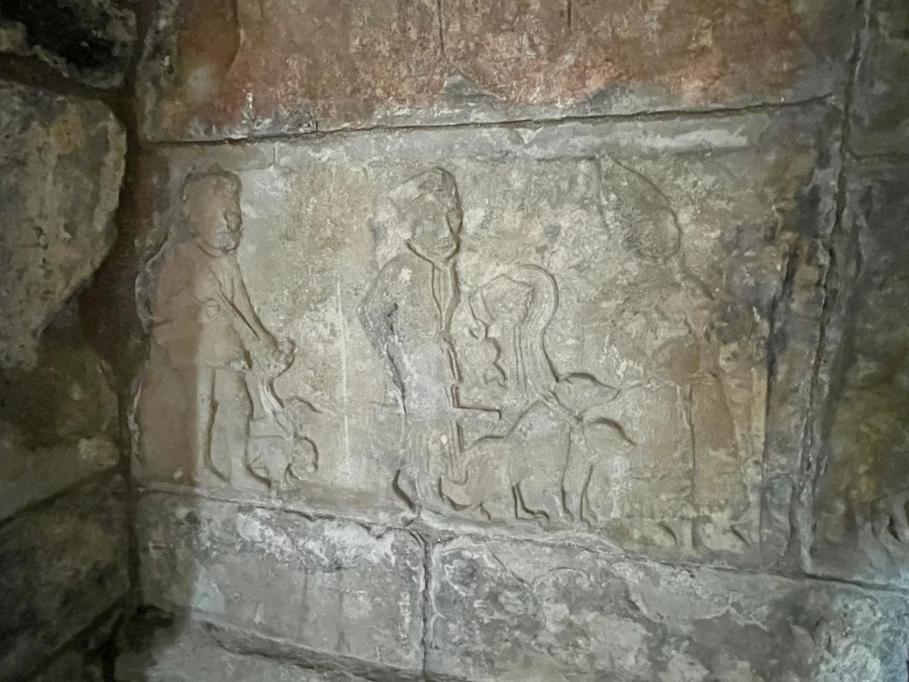 Flachrelief in Stein, das mehrere stehende Figuren in historischer Gewandung, begleitet von Hunden, zeigt. Die Darstellung ist im naiven, mittelalterlichen Stil gehalten und wirkt wie eine Prozession oder eine zeremonielle Szene.