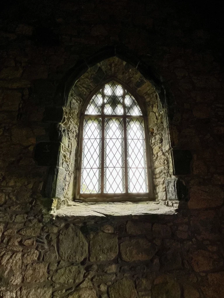 Ein spitzzulaufendes, gotisches Maßwerkfenster in einer dunklen Steinwand. Das Licht fällt durch die kleinteilige Bleiverglasung und erhellt die grobe Struktur der inneren Kirchenmauer.