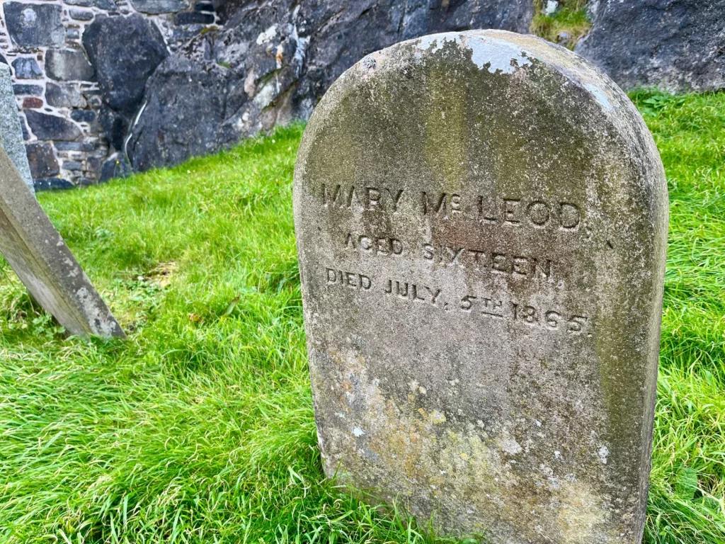 Eine detaillierte Frontalansicht eines verwitterten, abgerundeten Grabsteins. Die eingemeißelte Inschrift lautet: „MARY MCLEOD, AGED SIXTEEN, DIED JULY 5TH 1865“. Im Hintergrund ist eine grobe Steinmauer zu sehen.