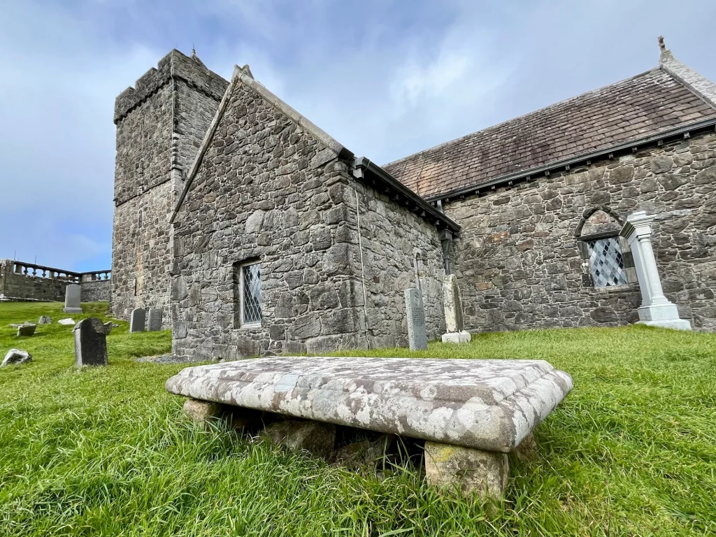Eine Froschperspektive auf die Steinkirche, im Vordergrund ein massiver, flacher Steintisch (ein sogenanntes Tischgrab), der auf kleinen Steinbeinen im Gras ruht.