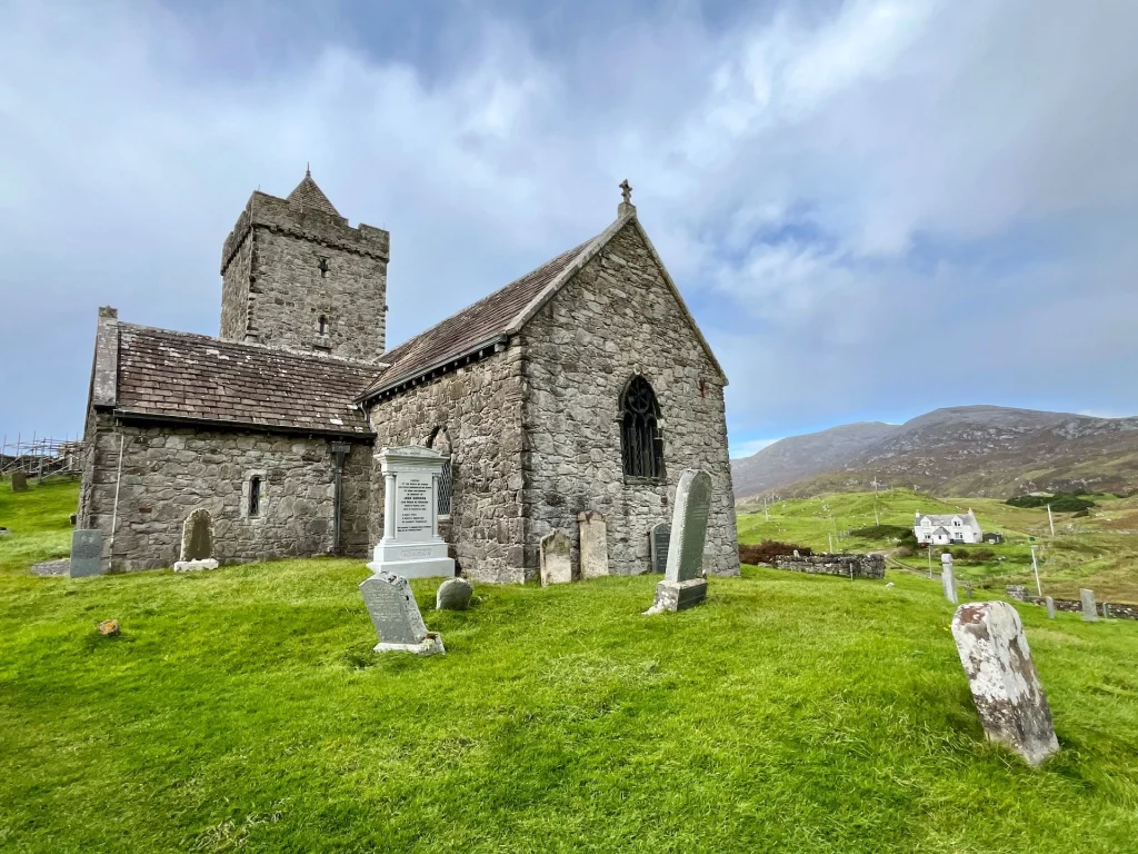 Eine Weitwinkelaufnahme der Kirche von der Seite, die das Zusammenspiel aus Querschiff, Hauptschiff und dem quadratischen Turm zeigt, umgeben von einem hügeligen Friedhof mit geneigten Grabsteinen.