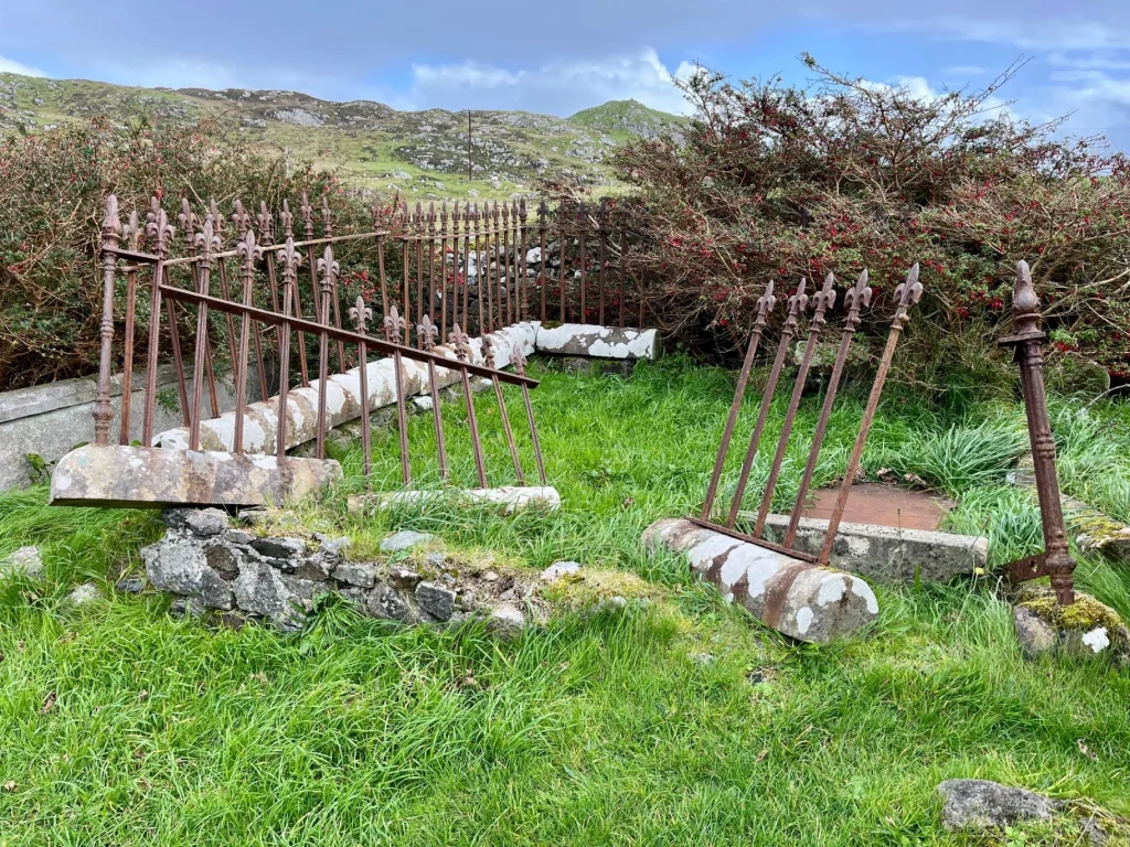 Nahaufnahme eines historischen, teils verfallenen eisernen Grabgeländers auf einem grasbewachsenen Friedhof. Die Metallstäbe sind verbogen und ruhen auf verwitterten Steinfundamenten vor struppigen Hecken.
