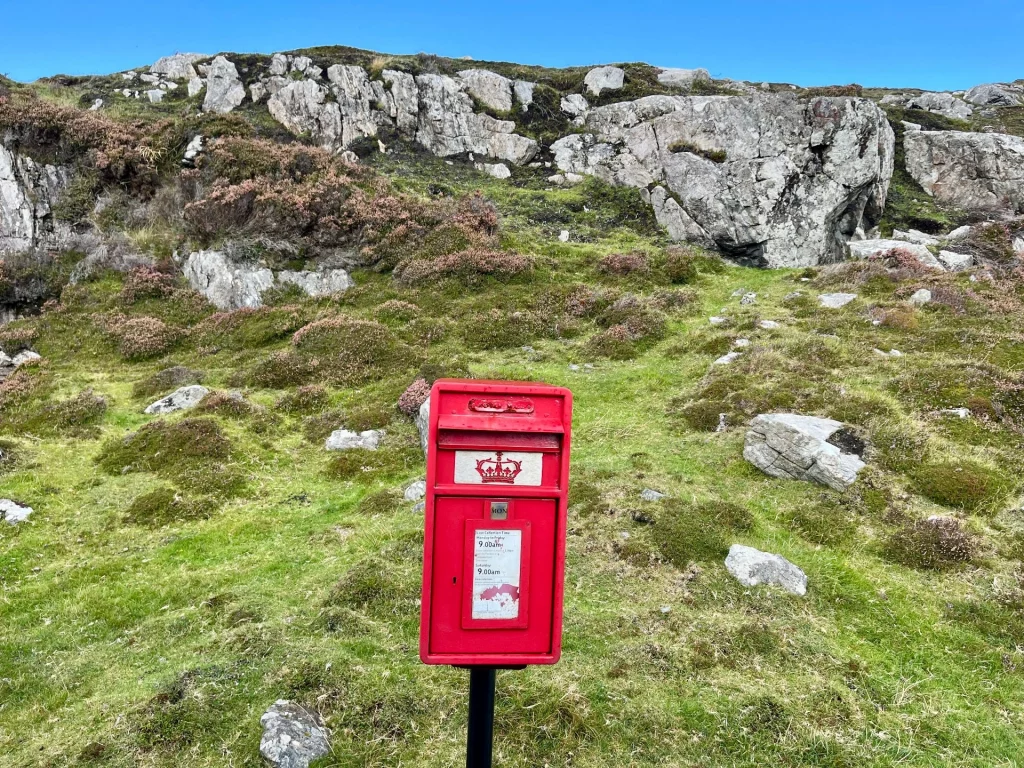Ein klassischer, knallroter britischer Postlaster (Postbox) steht einsam an einem grasbewachsenen Felshang unter freiem Himmel.