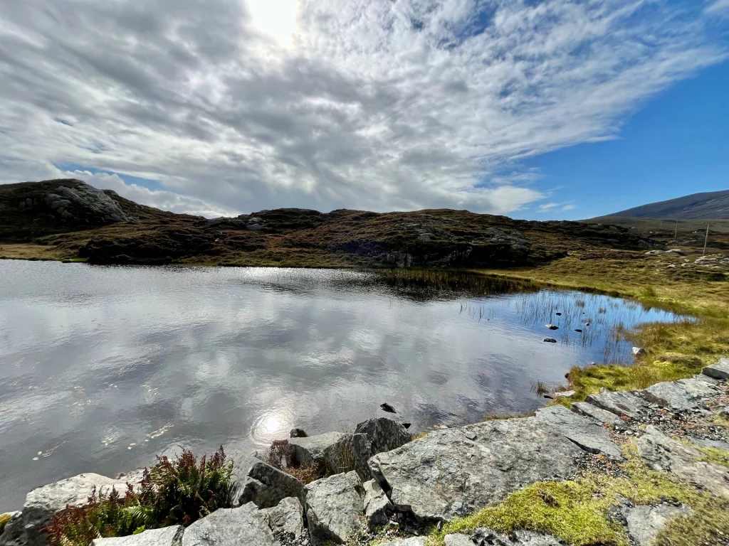 Ein wolkenverhangener Himmel spiegelt sich in der ruhigen Oberfläche eines kleinen Sees (Loch), dessen Ufer von grauen Felsen und dunklem Heidekraut gesäumt ist.