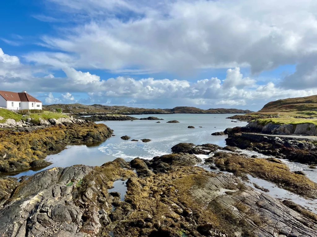Eine raue Küstenlandschaft mit dunklen Felsformationen und ruhigem Wasser unter einem weiten, bewölkten Himmel. Im Hintergrund sind vereinzelte weiße Häuser an der hügeligen Küstenlinie zu sehen.