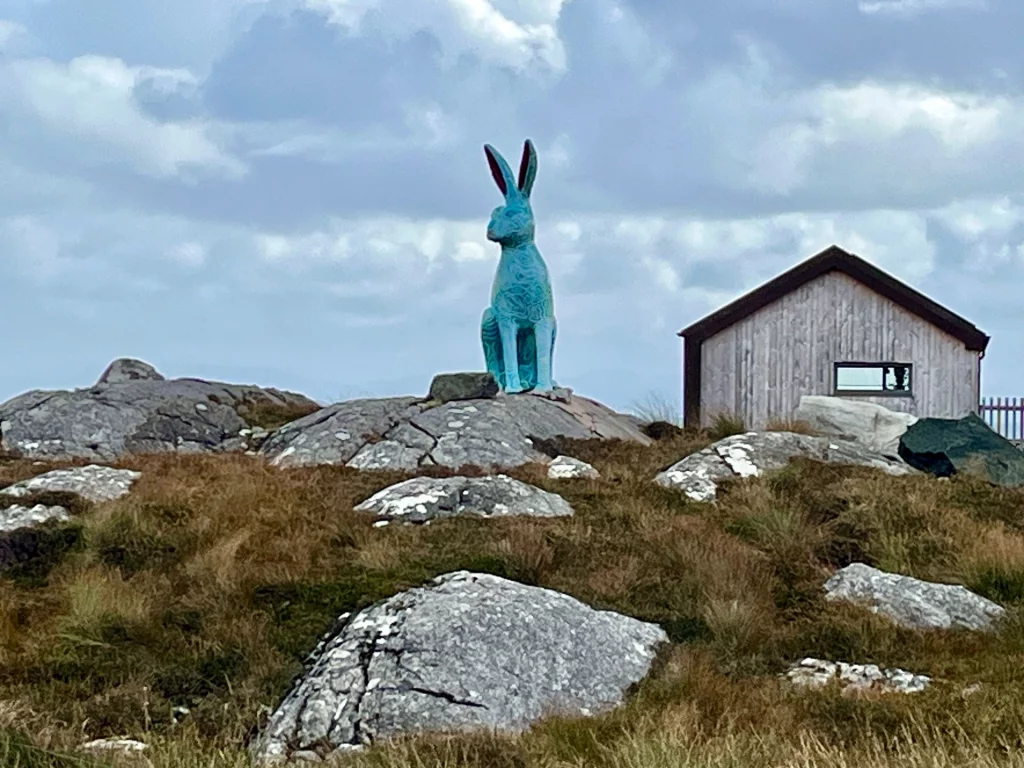 Eine auffällige, türkisfarbene Skulptur eines sitzenden Hasen auf einem felsigen Hügel neben einer kleinen Holzhütte. Die Skulptur bildet einen farbigen Kontrast zur natürlichen Umgebung.
