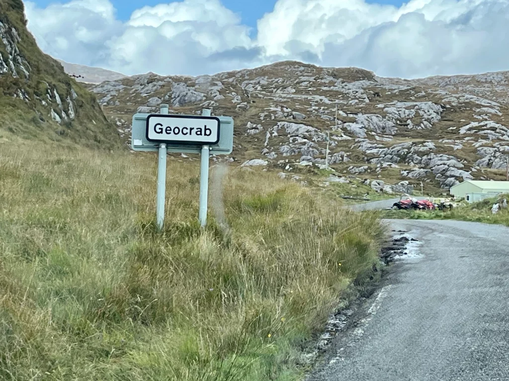Eine schmale, graue Asphaltstraße windet sich durch eine raue, felsige Landschaft mit trockenem Gras. Auf der linken Seite steht ein weißes Ortsschild mit der schwarzen Aufschrift „Geocrab“. Im Hintergrund ragen graue, zerklüftete Hügel unter einem dramatisch bewölkten Himmel auf.