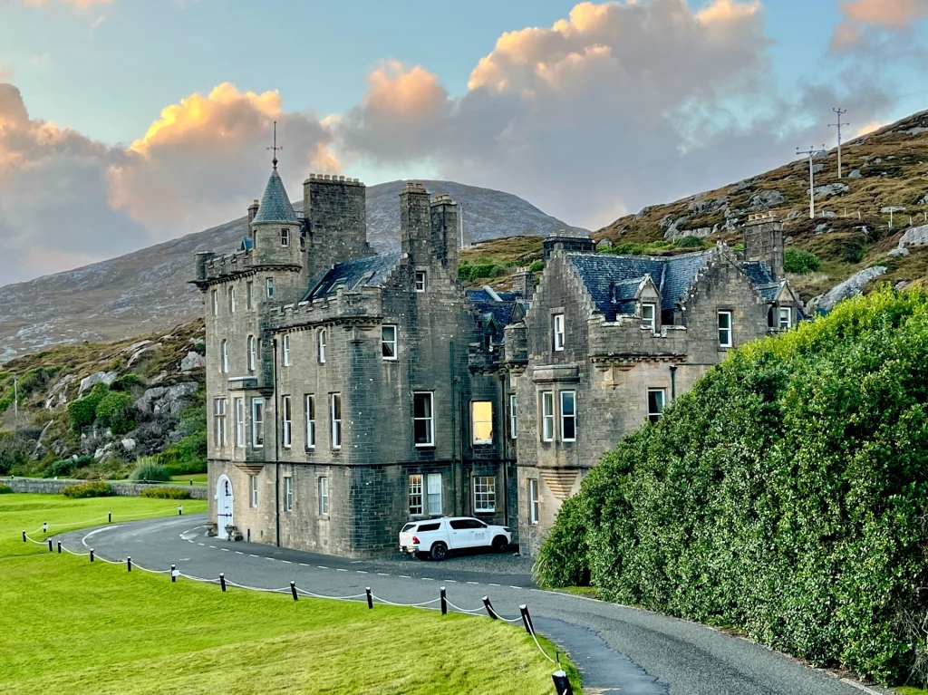 Ein großes, mehrstöckiges Herrenhaus aus grauem Naturstein im schottischen Baronialstil, gelegen vor einer kargen Hügellandschaft unter einem dramatischen Abendhimmel. Das Gebäude hat markante Türmchen, Giebel und zahlreiche Fenster. Ein weißer Pickup parkt in einer Nische des Hauses, während im Vordergrund eine gepflegte Rasenfläche und eine asphaltierte Zufahrt zu sehen sind.