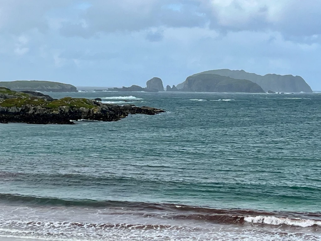 Eine raue Küstenszenerie mit türkisfarbenem Wasser und schäumender Brandung vor einer zerklüfteten Insel im Hintergrund unter bedecktem Himmel.