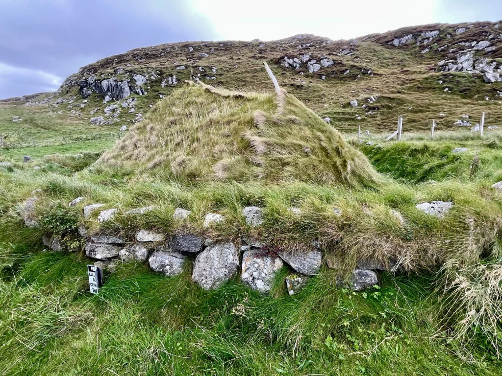 Rekonstruiertes Erdhaus: Ein mit Gras bewachsener Hügel, der auf einem Fundament aus Trockenmauerstein ruht – die Außenansicht eines Eisenzeithauses bei Bosta.