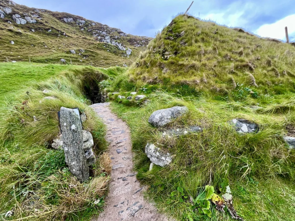 Ein schmaler, gepflasterter Steinweg führt zwischen grasbewachsenen Erdhügeln und Felsen direkt auf den dunklen Eingang einer prähistorischen Behausung zu.
