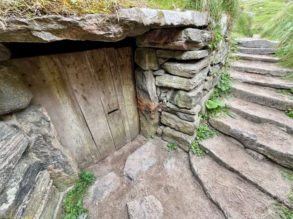 Eine niedrige, hölzerne Tür, die in einen Durchgang aus massiven, grob behauenen Natursteinen führt, die tief in den Boden eingelassen sind.