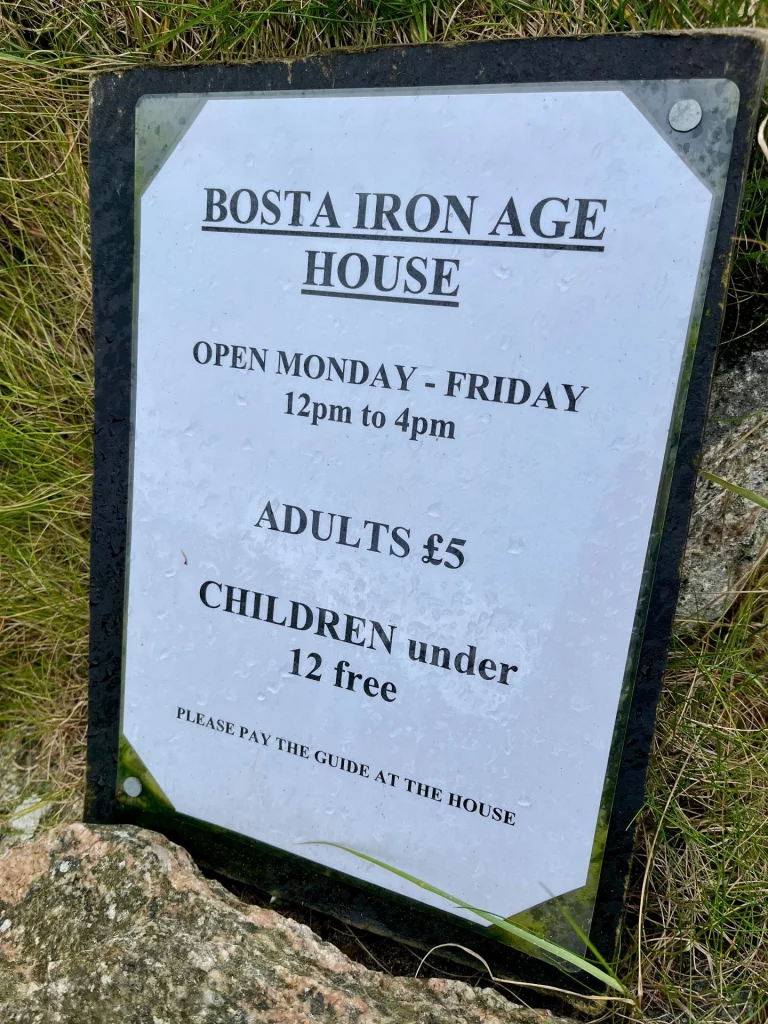 Ein Schild mit der Aufschrift „Bosta Iron Age House“, das die Öffnungszeiten und Eintrittspreise für Besucher ankündigt.