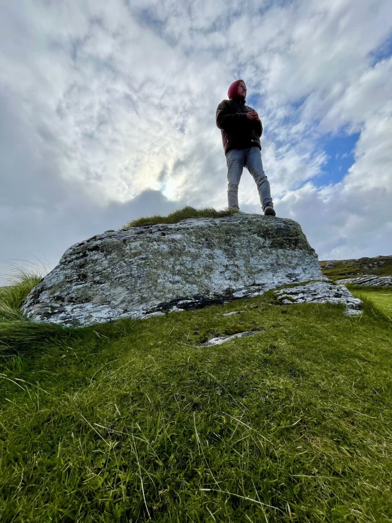 Ingo steht mit dem Rücken zum Betrachter auf einem großen, perfekt abgerundeten grauen Felsbrocken und blickt über eine weite, raue Moorlandschaft bis hin zum fernen Meer.