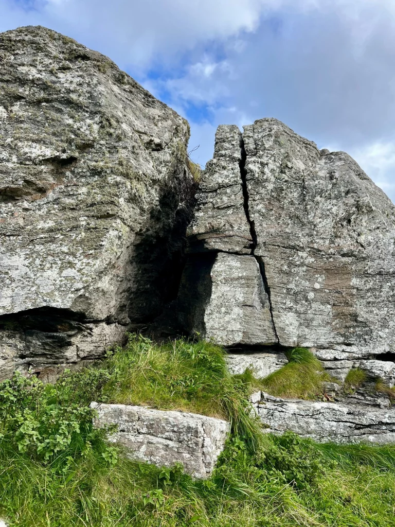 Ein massiver, aufrechter Felsblock, der in der Mitte durch einen sauberen, vertikalen Spalt geteilt ist, steht imposant in einer kargen, grünen Hügellandschaft.