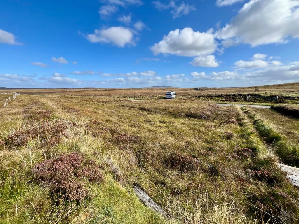 Blick über eine weite, herbstlich braune Heidelandschaft mit einem kleinen Feldweg und einem am Horizont parkenden hellgrauen Camper-Van.
