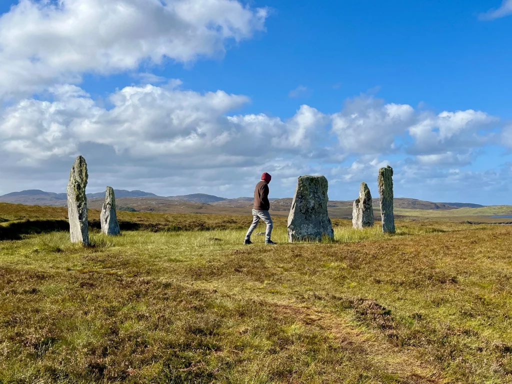 Eine weitläufige Aufnahme eines prähistorischen Steinkreises in einer Moorlandschaft, in dessen Mitte eine Person spaziert.