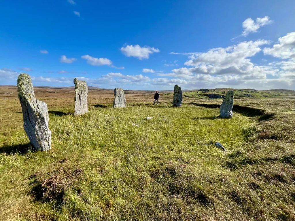 Ein Kreis aus grauen, hoch aufragenden Monolithen auf einem grünen Hügel unter einem wechselhaften schottischen Himmel.