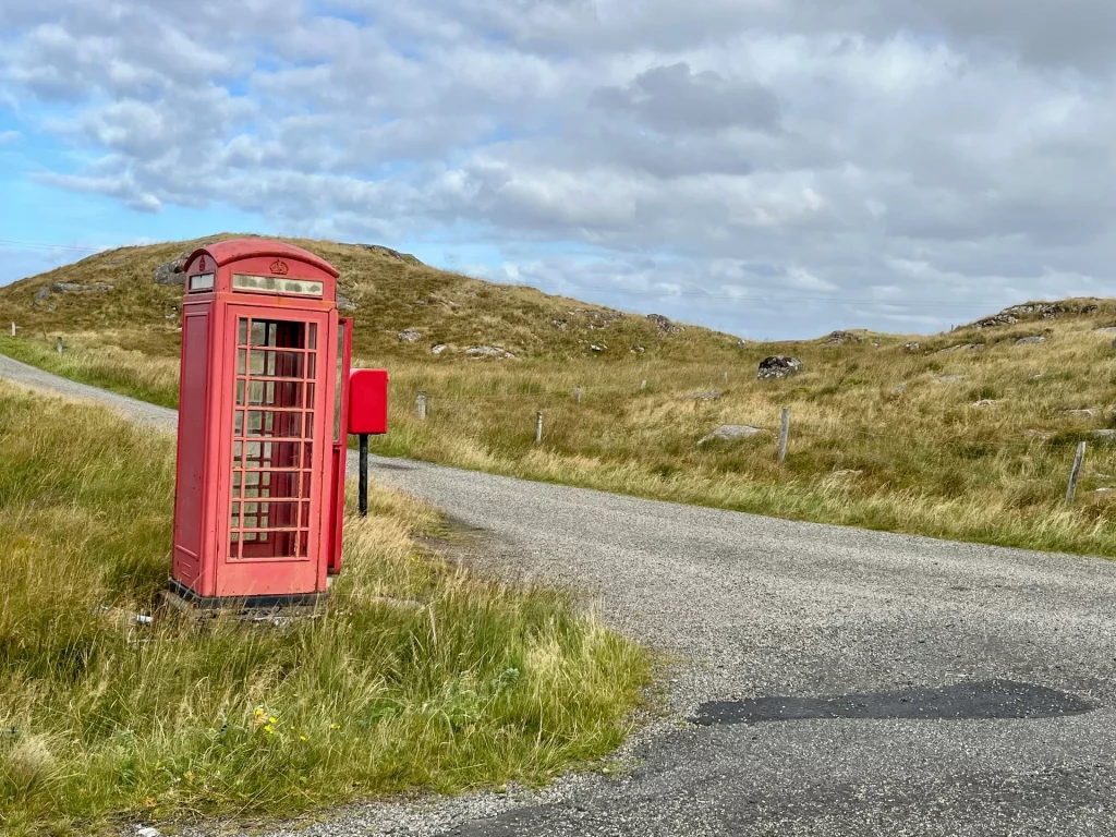 Eine klassische, rote britische Telefonzelle und ein kleiner Briefkasten stehen verlassen an einer Weggabelung in einer kargen, hügeligen Graslandschaft unter bewölktem Himmel.