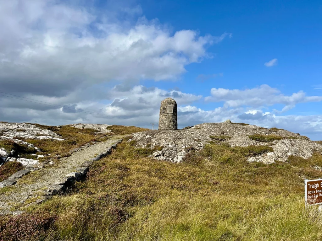 Ein schmaler, steiniger Pfad führt zu einem kleinen, runden Steinturm auf einer felsigen Anhöhe vor einem weiten Panorama.