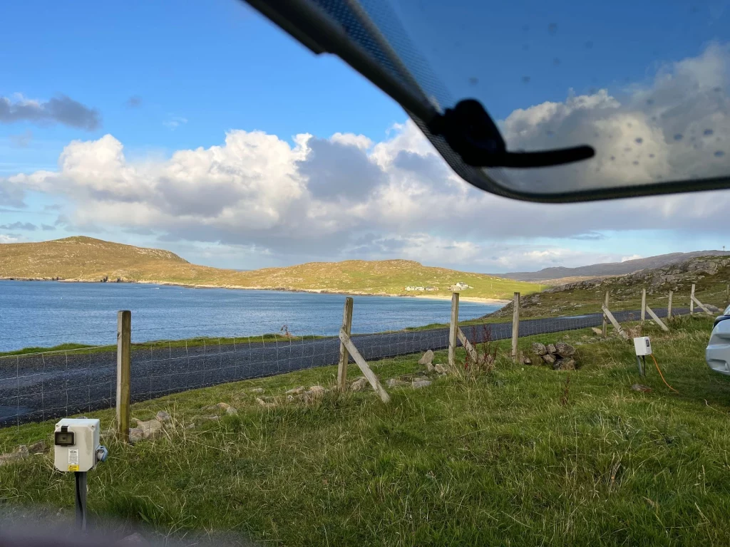 Blick aus einem Camper-Fenster auf eine Meeresbucht unter einem weitläufigen Wolkenhimmel. Im Vordergrund sieht man eine grüne Wiese mit einem Elektro-Anschluss für Wohnmobile und einen Holzzaun, der an einer asphaltierten Küstenstraße entlangführt. Im Hintergrund erstrecken sich sanfte, grüne Hügelketten bis zum blauen Wasser.