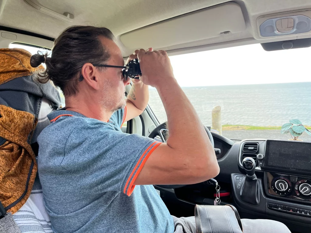Ingo sitzt konzentriert auf dem Fahrersitz der „Kätter“ und blickt durch ein großes schwarzes Fernglas aus dem Seitenfenster, um in der rauen schottischen Landschaft Adler zu entdecken - oder Robben.