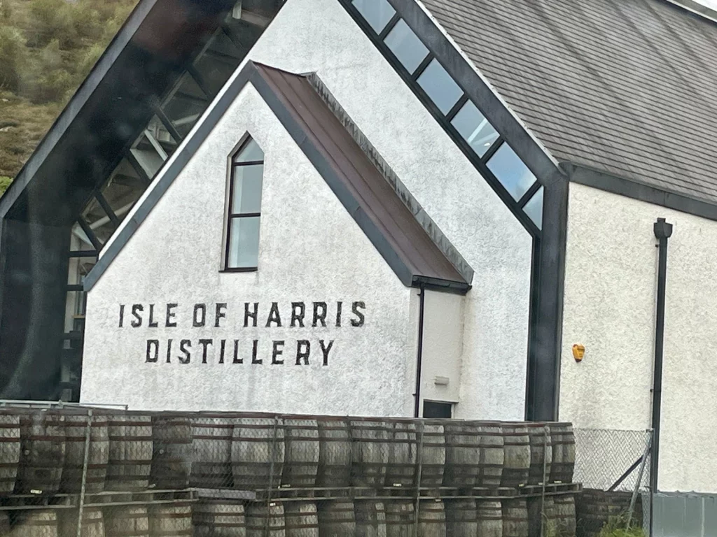 Außenansicht der Isle of Harris Distillery in Tarbert, Schottland. Das weiße Gebäude mit modernem Giebel zeigt den markanten Schriftzug der Brennerei. Im Vordergrund sind zahlreiche gestapelte Eichenfässer (Whiskyfässer) hinter einem Zaun gelagert.