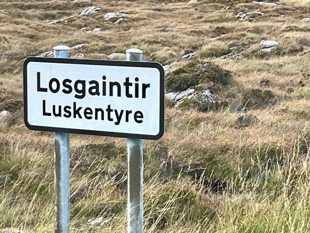 Ein zweisprachiges Ortsschild (Gälisch/Englisch) für „Losgaintir / Luskentyre“ vor der typisch schottischen Graslandschaft.