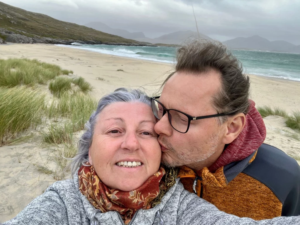 Ein glückliches Paar macht ein Selfie am windigen Strand; ein Mann gibt einer lächelnden Frau einen Kuss auf die Wange.