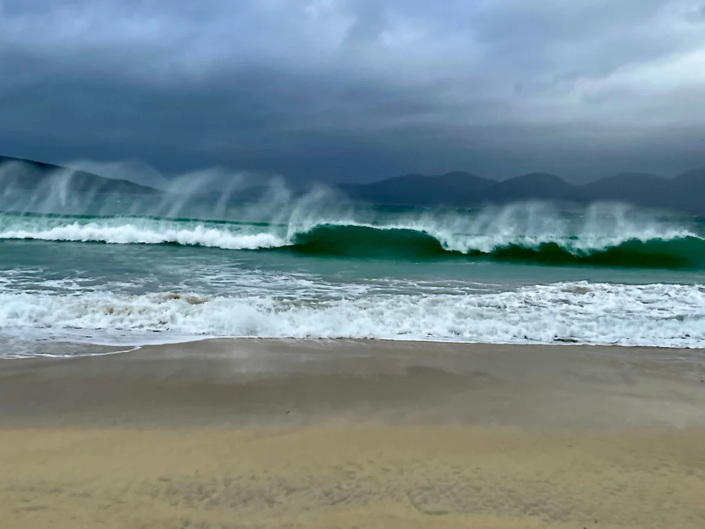 Große, smaragdgrüne Welle bricht am Ufer, während der Wind die Gischt über das Meer peitscht.