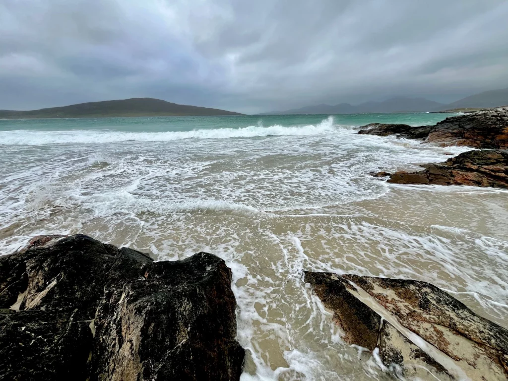 Dramatische Küstenlandschaft mit dunklen Felsen im Vordergrund, schäumenden Meereswellen und einer hügeligen Insel unter einem bedeckten, grauen Himmel.