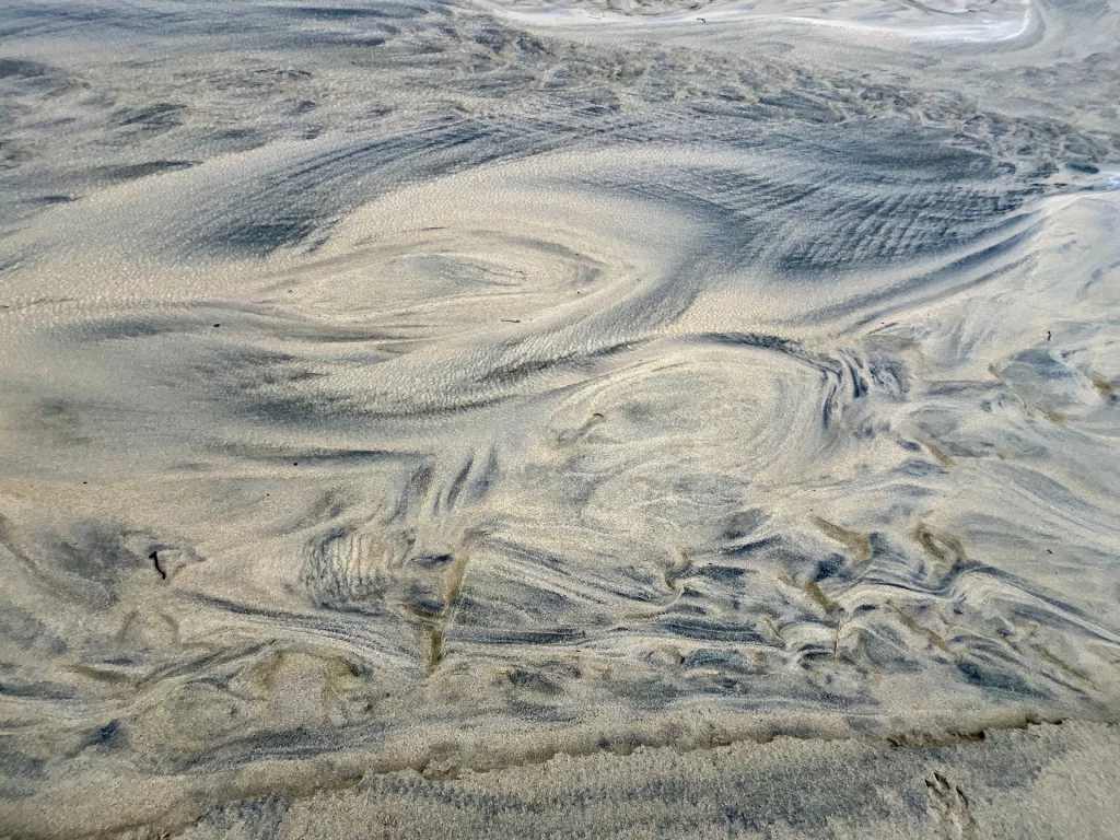 Abstrakte, kreisförmige Muster im feuchten Sand, die wie natürliche Marmorierungen am Strand der Hebriden wirken.