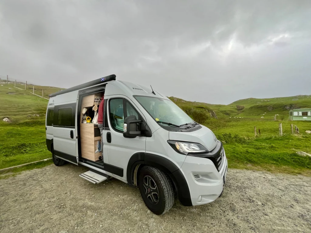 Ein hellgrauer Kastenwagen-Camper mit offener Schiebetür steht auf einem Schotterplatz in der grünen Hügellandschaft der Isle of Harris unter einem bewölkten schottischen Himmel.