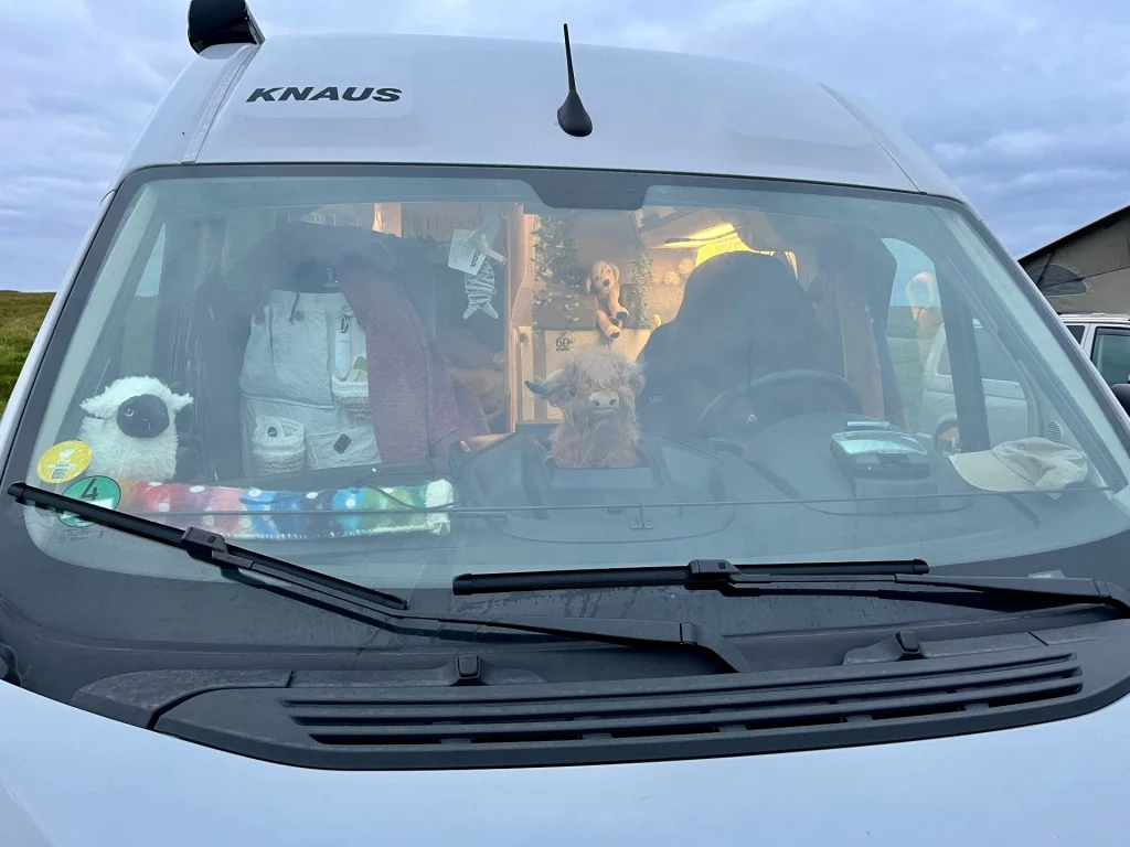 Blick durch die Frontscheibe eines weißen Knaus-Wohnmobils. Auf dem Armaturenbrett thronen ein plüschiges Highland-Rind und ein weißes Schaf als Maskottchen. Im gemütlich beleuchteten Innenraum sind weitere Kuscheltiere und Reiseutensilien zu erkennen.