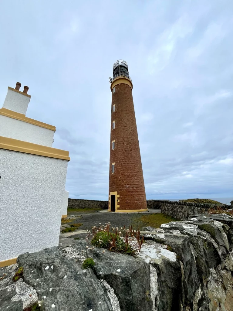 Ein hoher, runder Leuchtturm aus roten Backsteinen ragt unter einem bewölkten Himmel empor. Im Vordergrund sieht man eine rustikale Steinmauer mit kleinen Küstenpflanzen und den weißen Giebel eines Nebengebäudes mit markanten Schornsteinen.